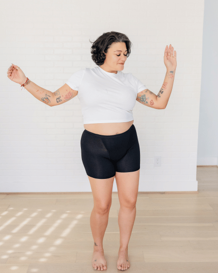 Person wearing a white crop top and black shorts standing with arms outstretched in a well-lit room.
